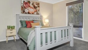 Ein Schlafzimmer mit einem weißen Bett, grünen und roten Kissen, einem Nachttisch mit Lampe und einem großen Blumenbild an der Wand.