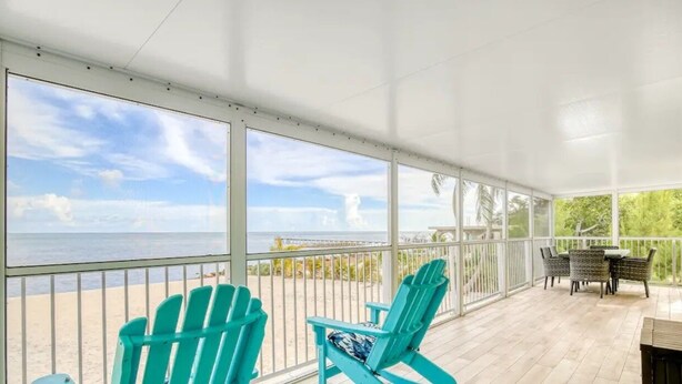 A covered outdoor area with a view of the beach, featuring two turquoise Adirondack chairs and a table with chairs.