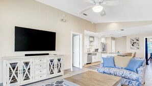 A spacious living room with a flat-screen TV, a white cabinet with drawers, a patterned sofa, and a ceiling fan.