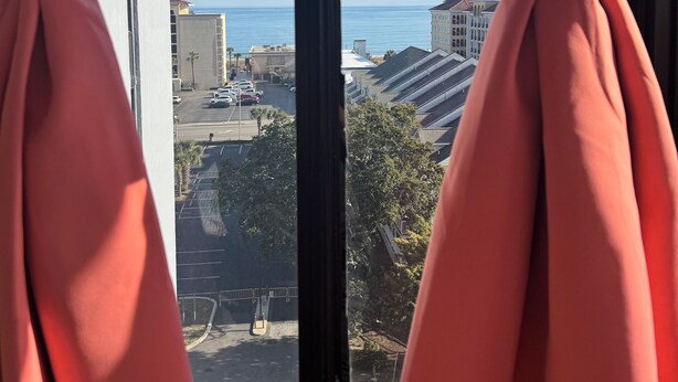 Ventana con cortinas rosadas atadas en la parte superior, con vista a un estacionamiento y edificios bajo un cielo azul despejado.