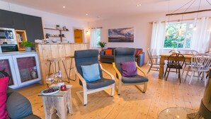A modern living room with a wooden floor, two grey armchairs, a small wooden table with wine glasses, and a dining area with a wooden table and chairs. - Eigionfa -  an on the beach that sleeps 8 guests  in 3 bedrooms (Menai Bridge)