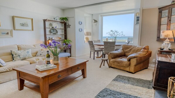 Una sala de estar con sofá, mesa de centro y sillón de cuero. Se observa una mesa de comedor con sillas y vista al mar a través de un ventanal grande.