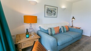 Una sala de estar acogedora con un sofá azul, una mesita de madera con una botella de vino y copas, una lámpara y un cuadro en la pared.