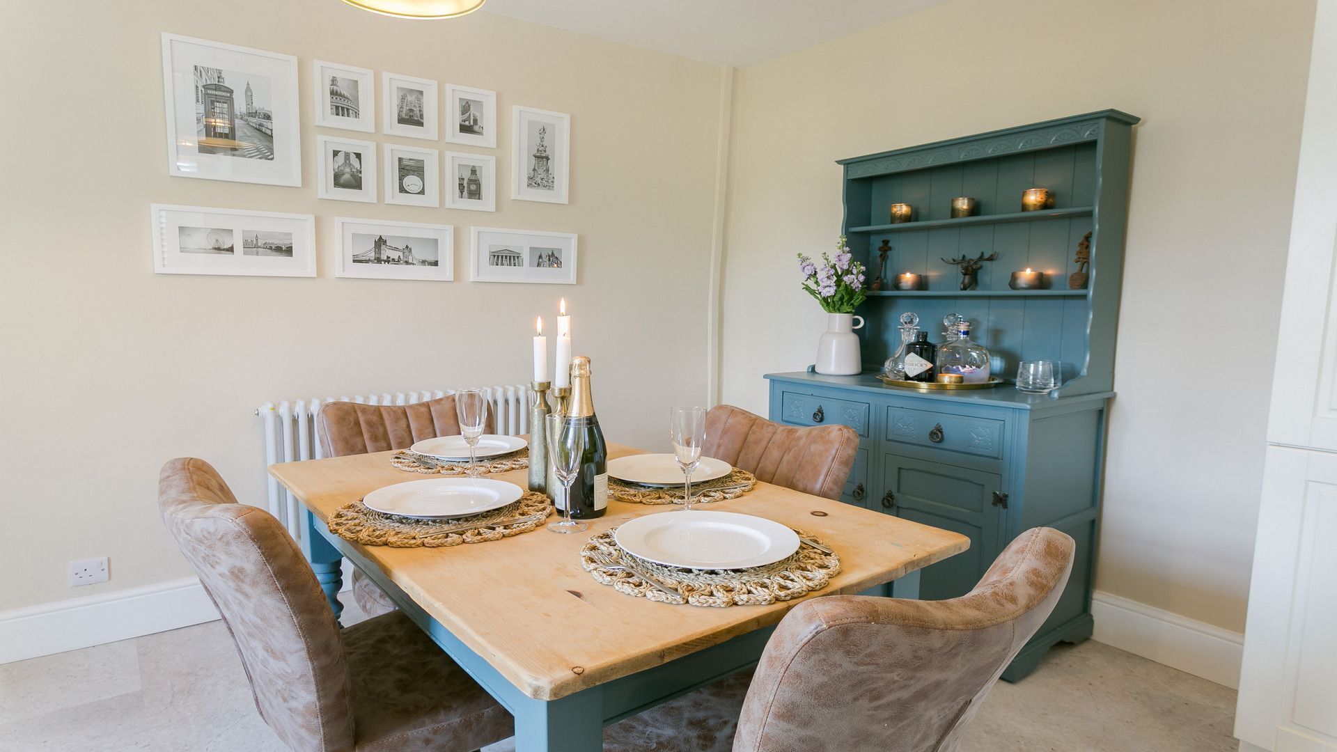 Une salle à manger avec une table en bois dressée pour quatre personnes, entourée de chaises rembourrées. Au-dessus de la table, des photographies en noir et blanc encadrées ornent le mur. À droite, on trouve un meuble de couleur turquoise, agrémenté d’objets décoratifs et de bougies.