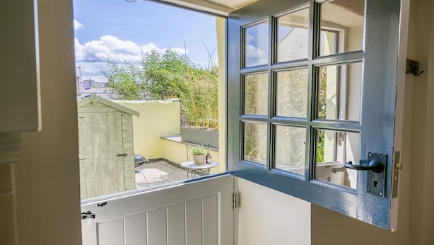 A view through a partially open door with a window, revealing a small outdoor area with a table, a potted plant, and a barbecue grill. - Ty Melyn -  sleeps 4 guests  in 2 bedrooms (Beaumaris)