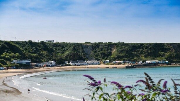 A coastal area with a sandy beach, a clear blue sea, and a hillside with buildings.