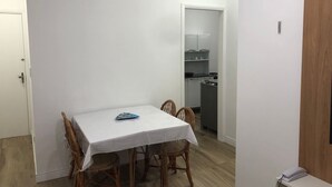Una habitación pequeña y limpia con una mesa de comedor, sillas de mimbre, una puerta blanca y una kitchenette al fondo.