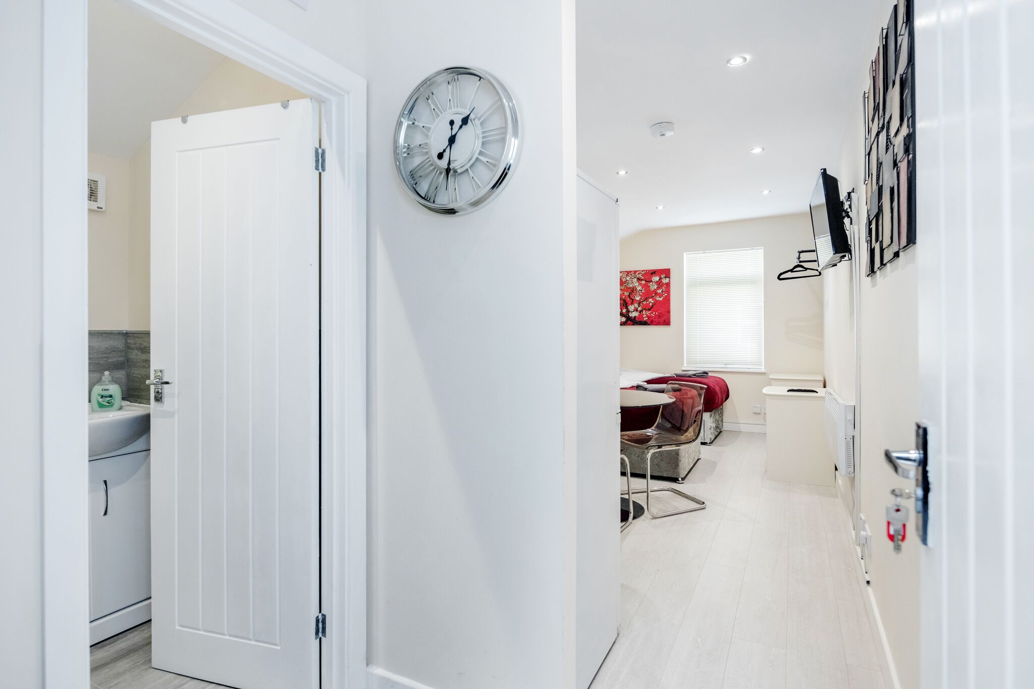 A modern kitchen with a white door, a wall-mounted clock, and a dining area with a red decorative piece.