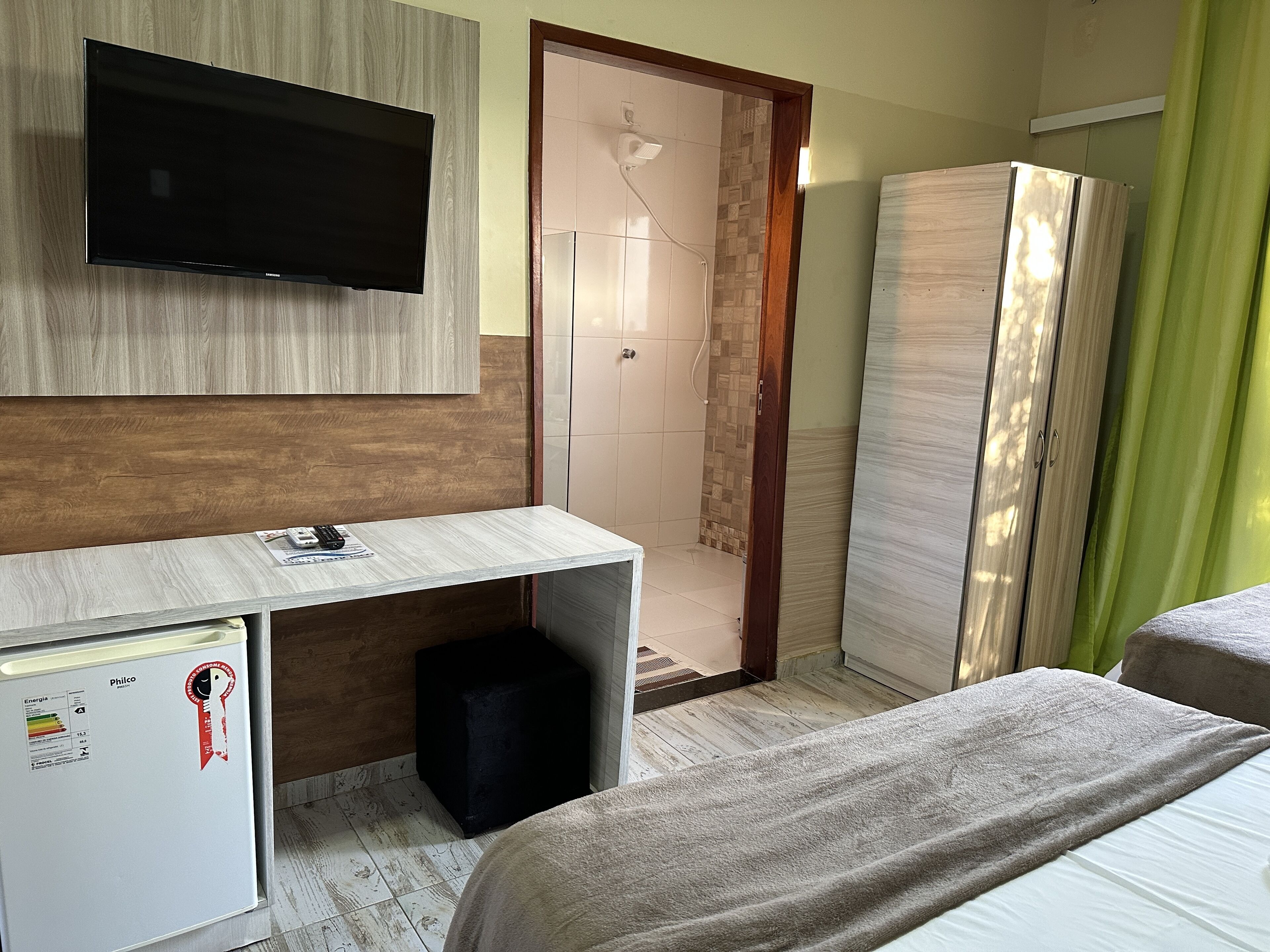 A hotel room with a flat-screen TV mounted on the wall, a desk with a computer and a small refrigerator, a walk-in closet, and a bed with a grey blanket.