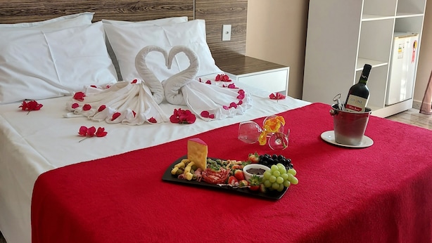 Una cama cuidadosamente arreglada con un edredón rojo, una almohada con forma de corazón y pétalos de rosa. Hay una bandeja con aperitivos y una botella de vino sobre la cama.