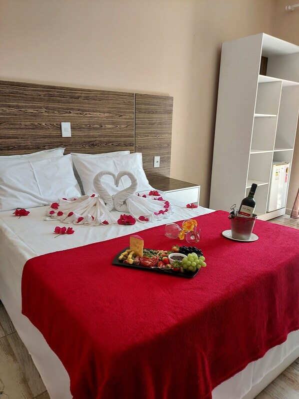 A neatly made bed with a red blanket, a heart-shaped pillow, and rose petals. There is a tray with snacks and a bottle of wine on the bed. - Hotel São Francisco (Piranhas)