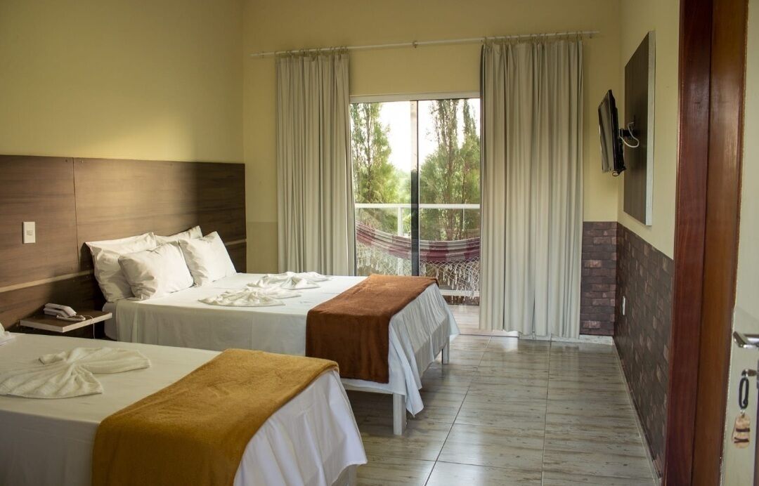 Quarto de hotel com duas camas, varanda com cortinas, televisão na parede e cabeceira de madeira.