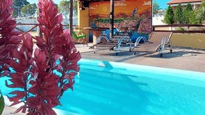Una piscina con sillas de descanso, un espacio cubierto con un mural, y un cielo azul despejado.