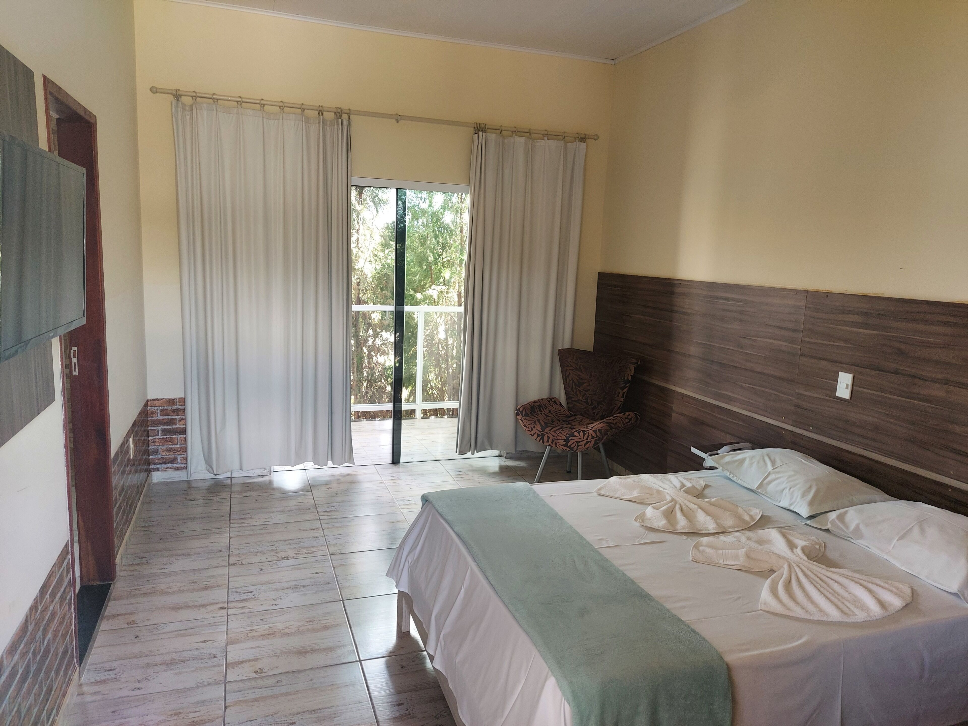 Habitación de hotel con cama, una silla, un ventanal con cortinas y vistas a zonas verdes.