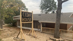 Et træskilt med teksten "CEDAR HOUSE" foran en bygning med en overdækket veranda og en grusvej.