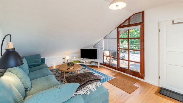 Een loft-woonkamer met schuin dak, een televisie op een witte standaard, een bank met kussens, een lamp en uitzicht door een groot raam.