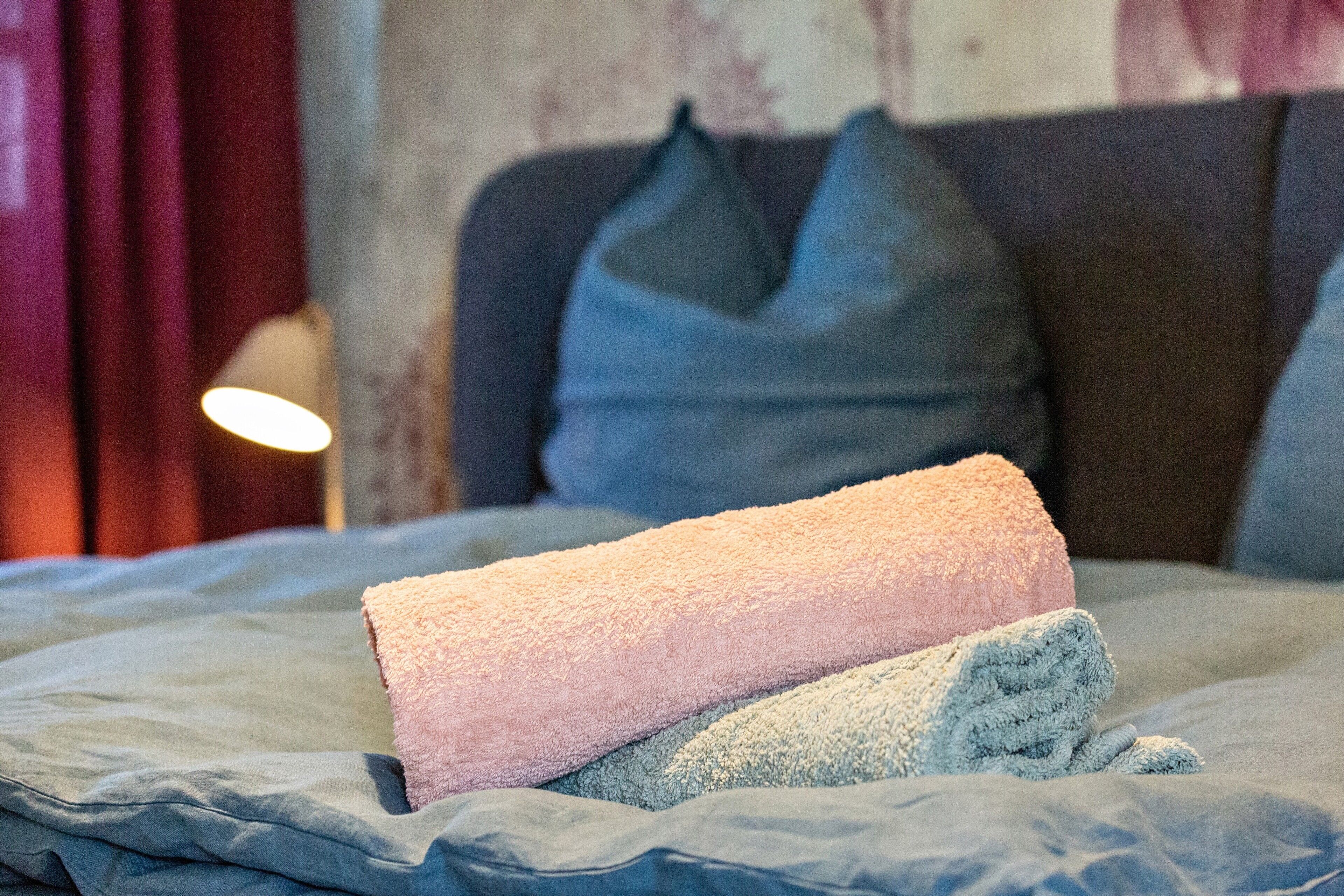 A bed with a rolled-up pink and blue towel, a dark headboard, and a lamp in the background.