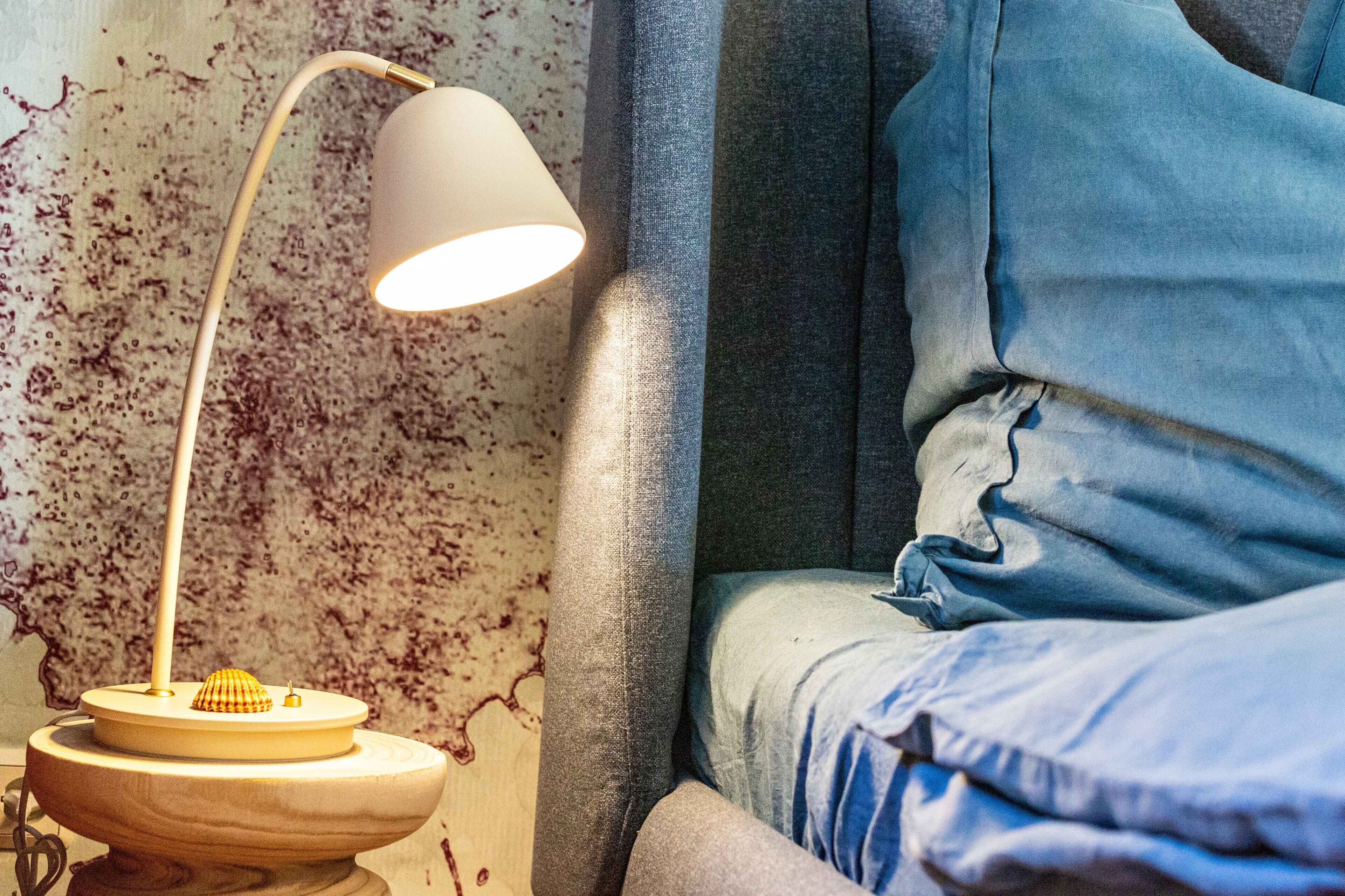 Een bed met een blauwe deken, een witte lamp op een houten voet en een wand met een puntpatroon.