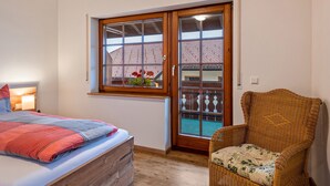 A bedroom with a bed, a wicker chair, a window with a view of a building, and a wooden door.