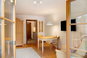 Een eetruimte met een houten tafel en stoelen, een wit vloerkleed en een aan de muur bevestigde televisie.