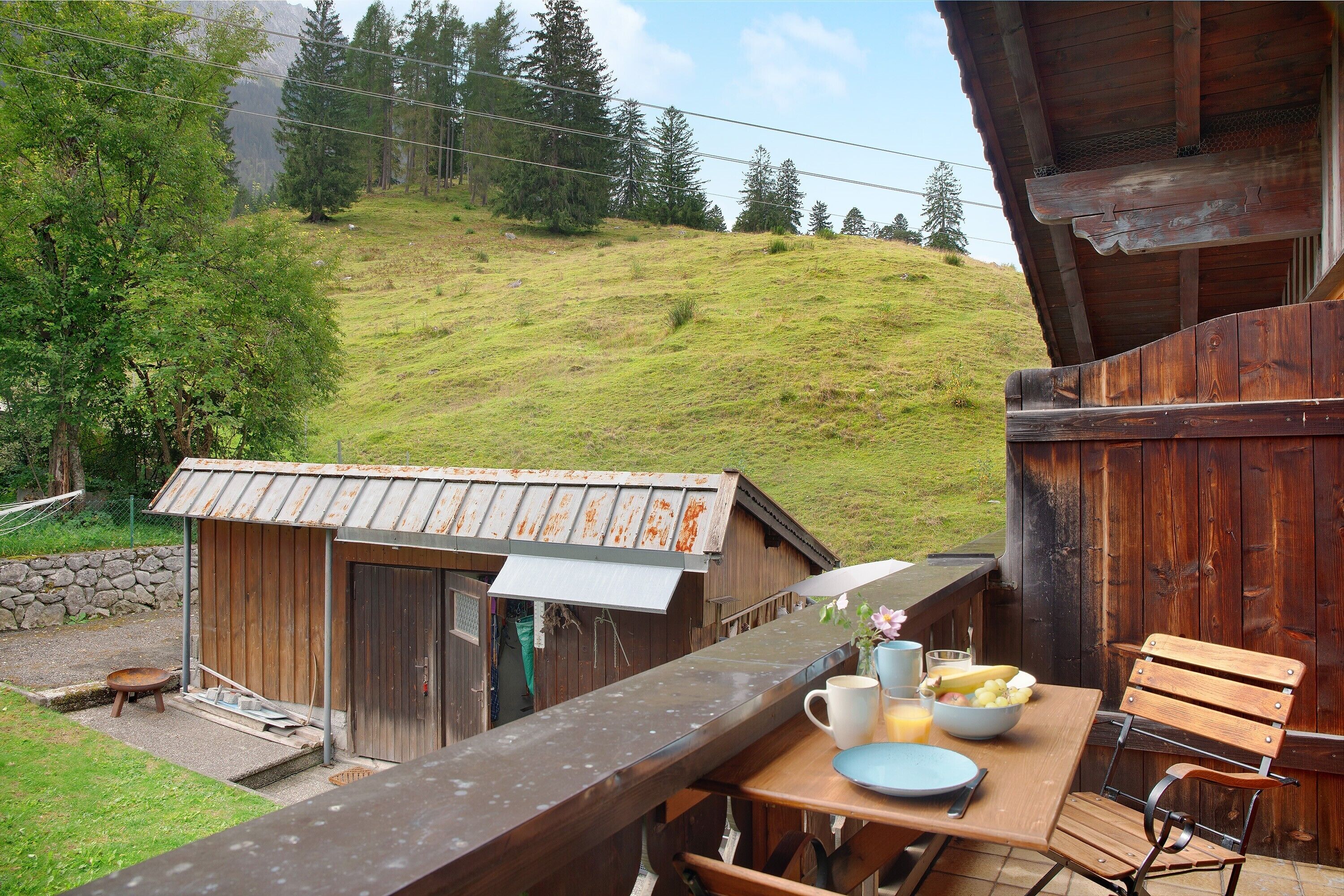 Eine Holzterrasse mit Tisch und Stühlen, eine kleine Holzhütte und ein grasbewachsener Hang im Hintergrund.