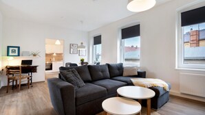 A modern living room with a grey sectional sofa, a small round table, a desk with a chair, and a view of the city through large windows. - Holiday Apartment "Finsterwalde" (FInsterwalde)