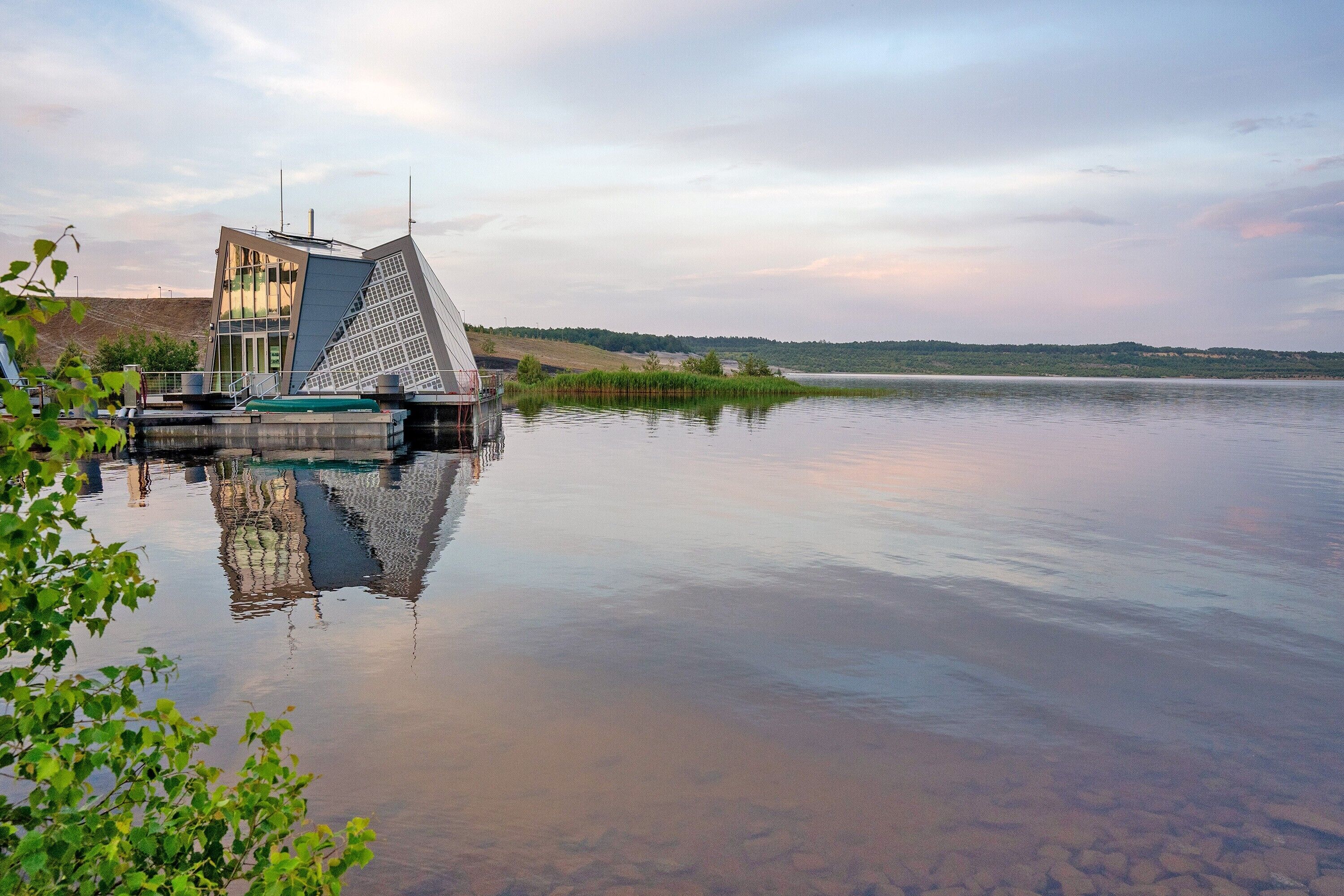 Ein modernes Gebäude mit einem markanten, dreieckigen Dach, einem Anleger und ruhigem Wasser, das die Architektur spiegelt.