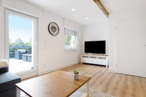 A modern living room with a wooden coffee table, a television on a white stand, and a large window with a view of outdoor furniture.
