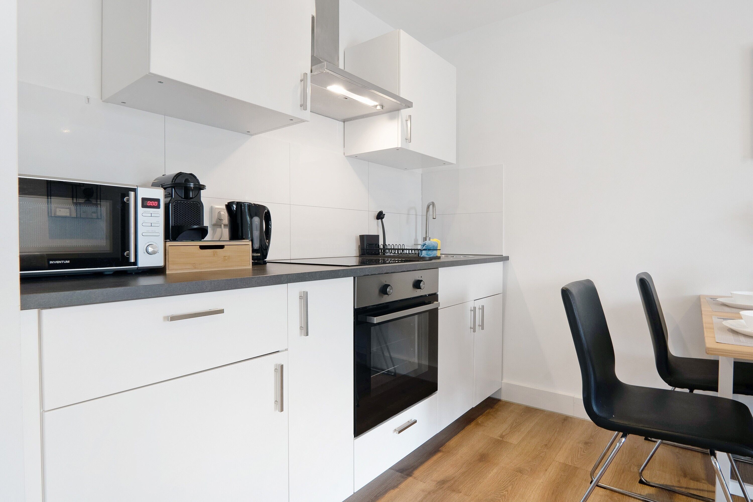 Una cocina moderna con armarios blancos, microondas, cafetera y comedor con sillas negras.