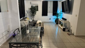 A modern living room with a glass dining table, black chairs, a sofa, a TV mounted on the wall, and a potted plant.