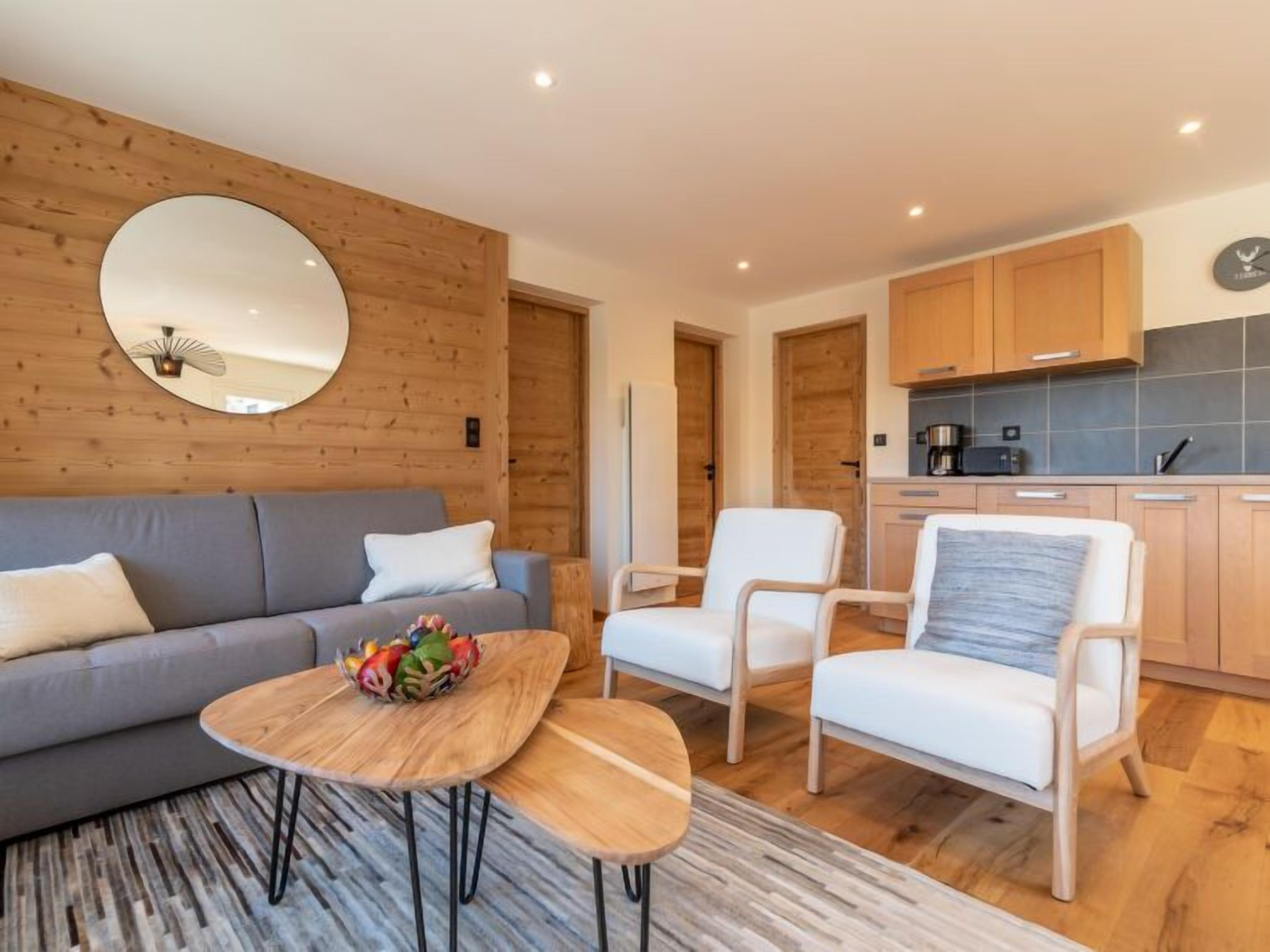Un salon moderne avec un canapé gris, des fauteuils blancs, une table basse en bois et un miroir rond accroché au mur.