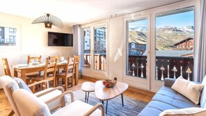 Una sala moderna con un sofá, sillones, una mesa de comedor y un televisor. Amplias ventanas ofrecen vistas a edificios y montañas.