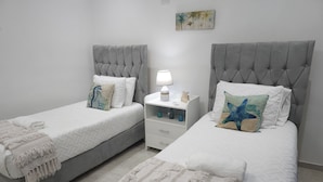 Une chambre à coucher avec deux lits, chacun doté d’une tête de lit grise, de draps blancs et de coussins décoratifs. On y trouve une table de chevet avec une lampe, ainsi qu’une petite étagère sur laquelle sont posés divers objets.