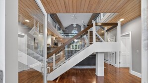 Un interior amplio con una escalera de madera, barandillas de vidrio y una cocina moderna al fondo.