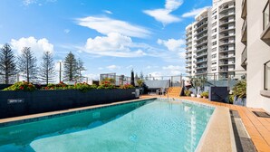 A swimming pool with a depth marker, surrounded by a landscaped area with plants and a view of a high-rise building.