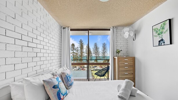 A bedroom with a white brick wall, a bed with white bedding, a wooden dresser, a fan, and a view of trees and a parking lot through a glass door.