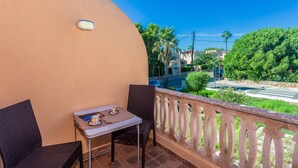 A balcony with two wicker chairs, a small table with a cup and saucer, and a view of a street with palm trees and houses. - Apartment 'Relax' with Shared Garden, Balcony and Wi-Fi (Ciudadela de Menorca)