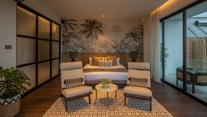 A modern bedroom with a large bed, two armchairs, a small table, and a view of the outside. - Mandala Clubhouse (Canggu)