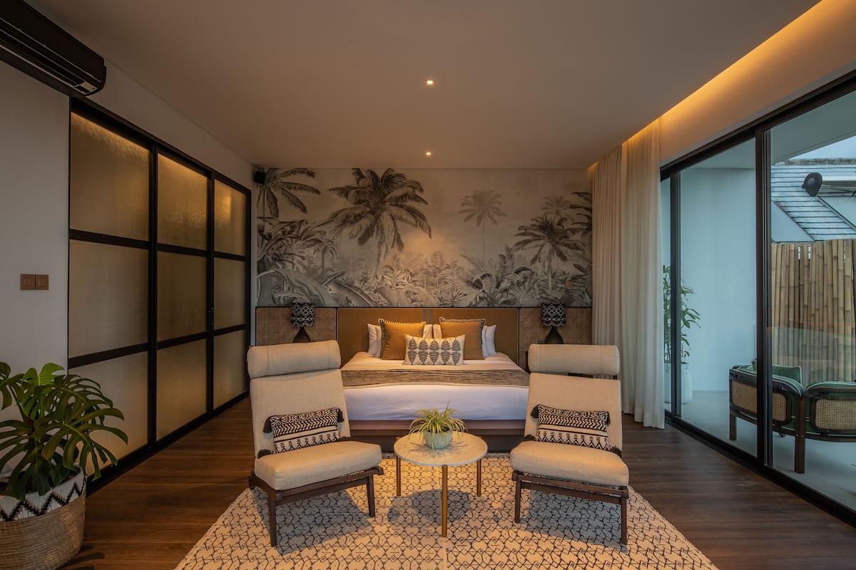 A modern bedroom with a large bed, two armchairs, a small table, and a view of the outside.