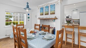 Un comedor con una mesa preparada para cuatro personas, sillas de madera y un mantel azul. Hay una zona de cocina con microondas y refrigerador.