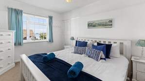 Un dormitorio con cama, cómoda, ventana con cortinas, lámpara de noche y un cuadro en la pared.