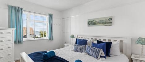 Ein Schlafzimmer mit Bett, Schminktisch, Fenster mit Vorhängen, Nachttischlampe und einem Gemälde an der Wand.