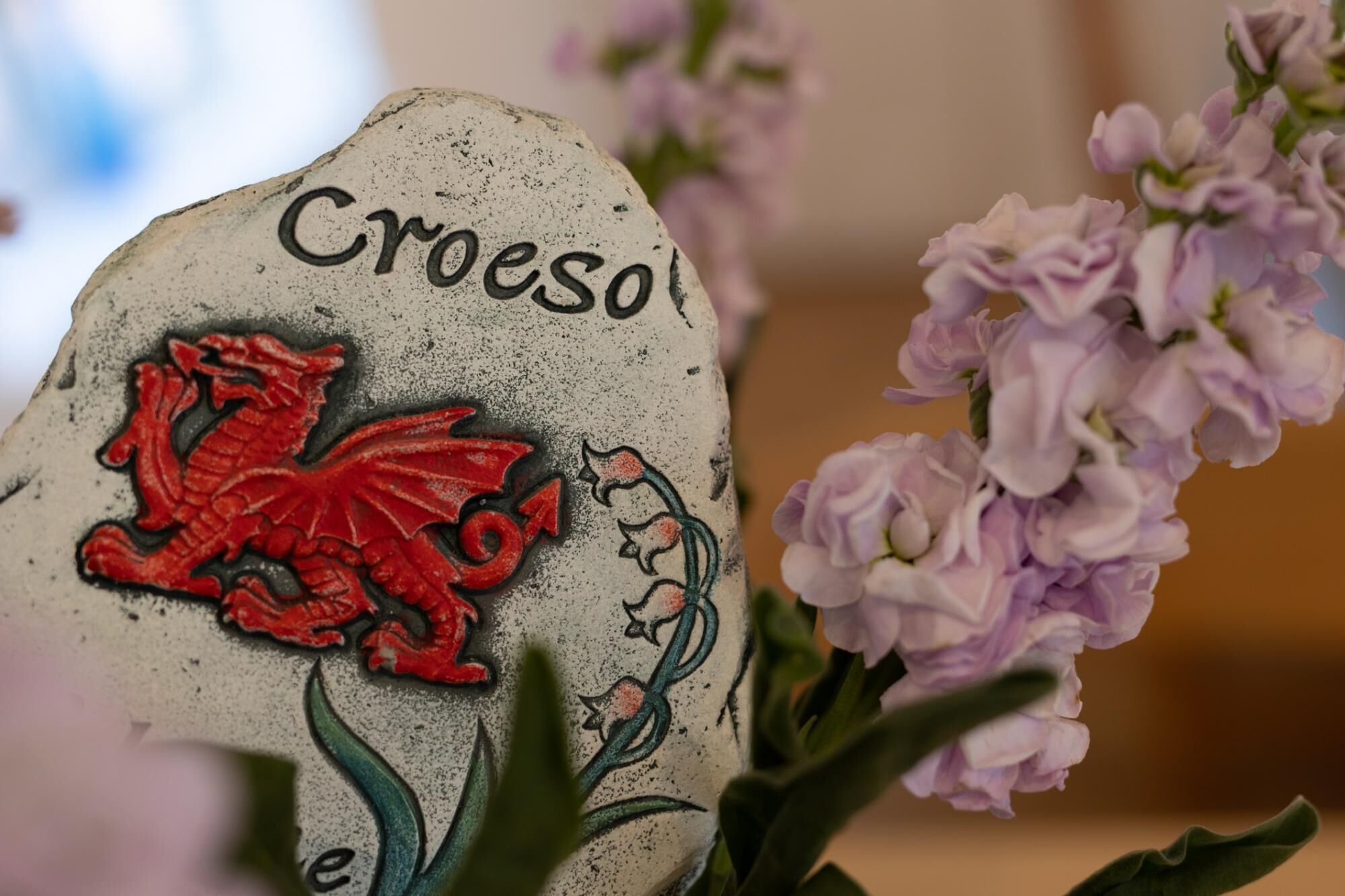 Une pierre sur laquelle est gravé le mot « Croeso » et un emblème représentant un dragon rouge, le tout entouré de fleurs violettes.