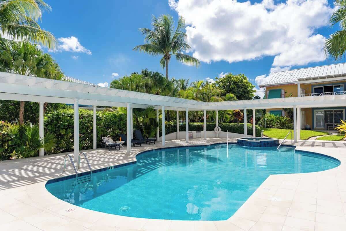 Une piscine agrémentée d’une pergola blanche, de palmiers et d’un bâtiment visible au fond.
