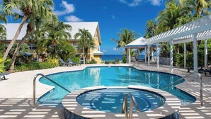 Une piscine avec un jacuzzi, entourée de palmiers et bordée d’un bâtiment au toit blanc.