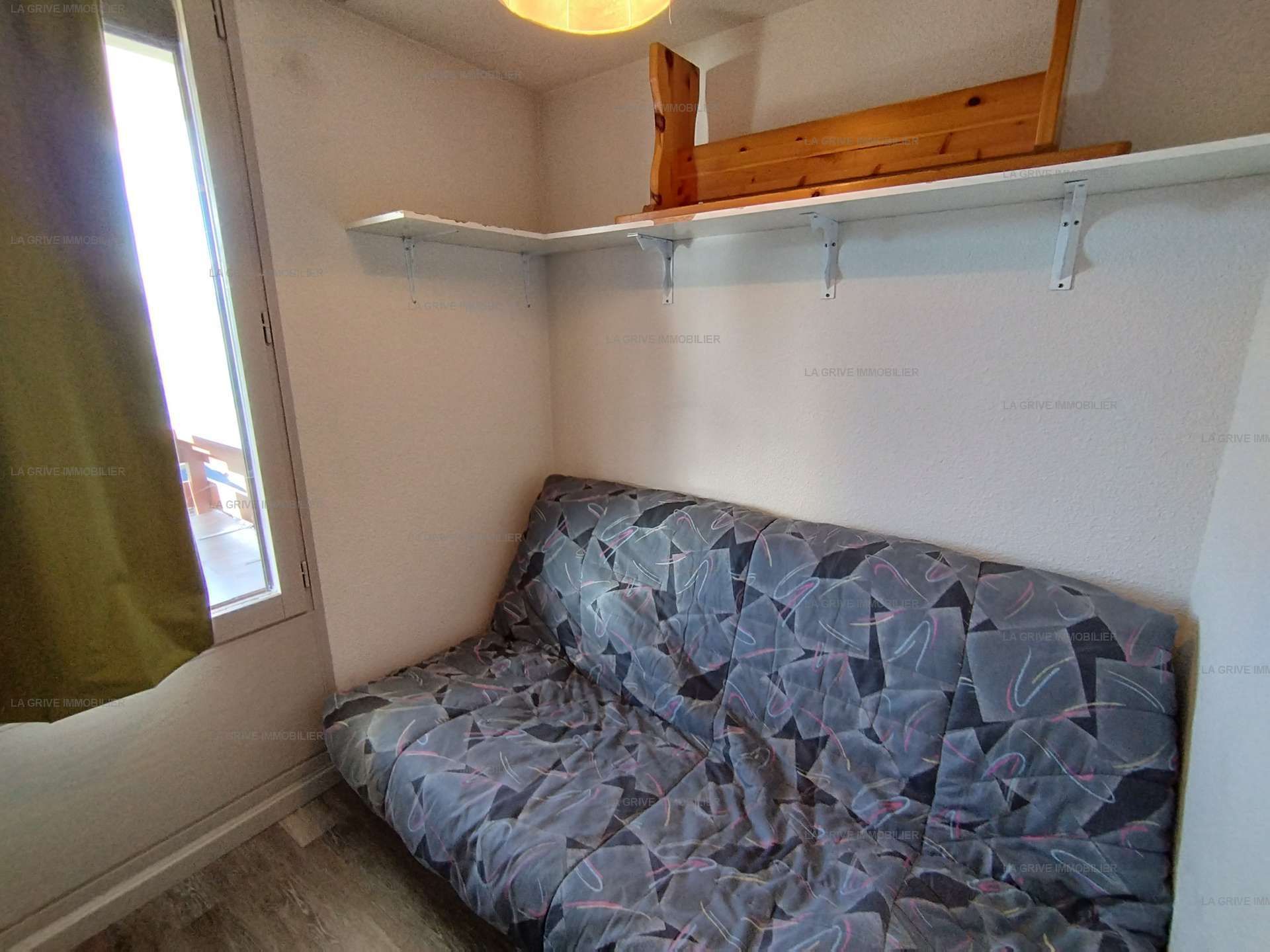 Une petite pièce avec un lit simple, une fenêtre et une étagère en bois.