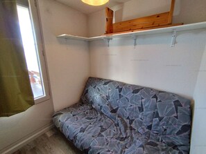 Ein kleines Zimmer mit einem Einzelbett, einem Fenster und einem Holzregal.