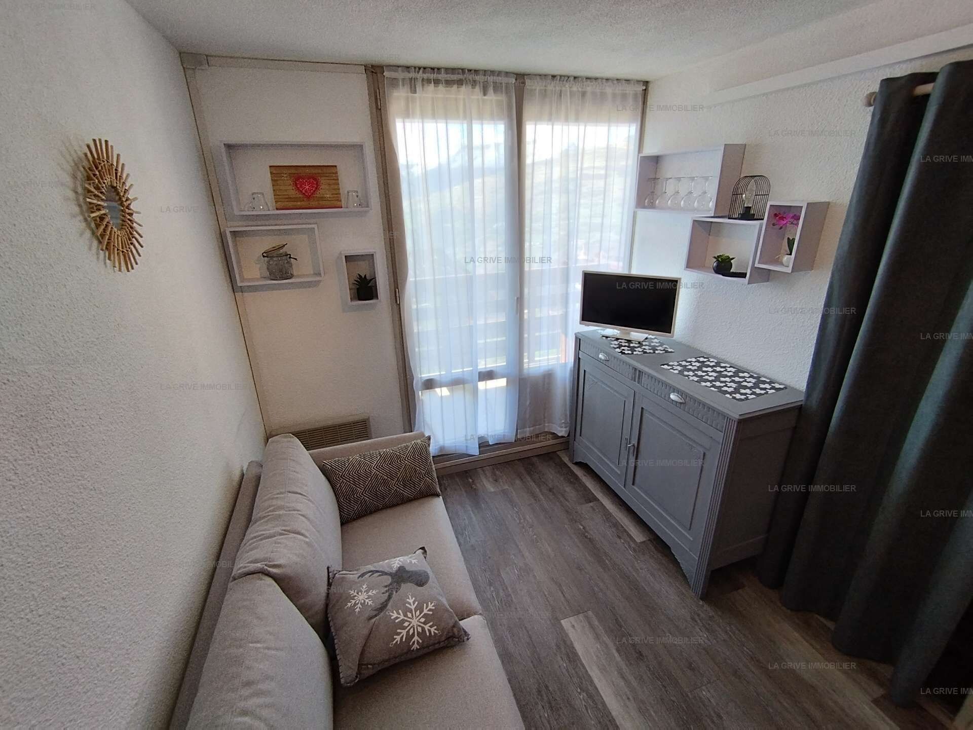 Un espace de vie compact comprenant un canapé, un téléviseur, un petit coin cuisine et une fenêtre agrémentée de rideaux légers.