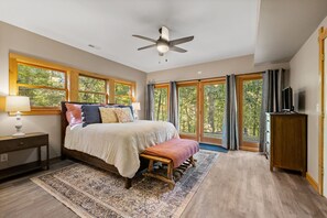 Une chambre avec un grand lit, un ventilateur de plafond, une table de chevet, un téléviseur et une vue sur les arbres par les fenêtres.
