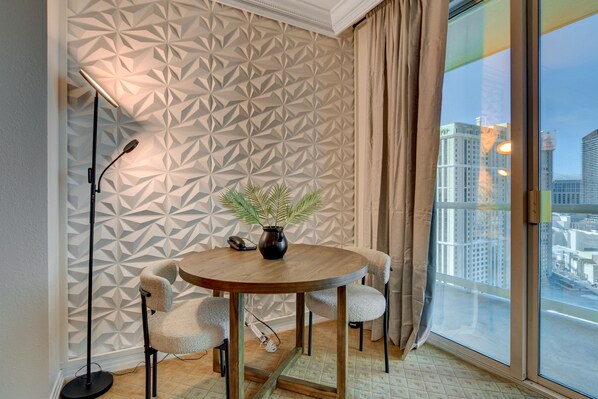 A modern room with a geometric patterned wall, a round wooden table with chairs, a floor lamp, and a view of a cityscape through a large window. - Studio 25 HIGH FLR MGM Signature Balcony (Las Vegas)